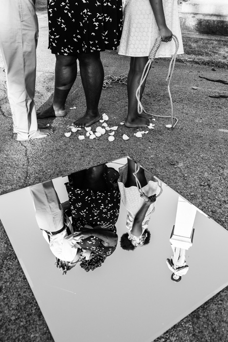 Two Black women kissing while a young Black girl stands with her back to them, holding a noose. The image reflected in a mirror on the ground also shows a confederate statue