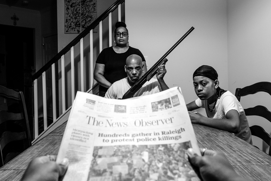 In the foreground, a newspaper with the headline "Hundreds gather in Raleigh to protest police killings." In the background, a Black woman, man and child surround a table; the man holds a shotgun.