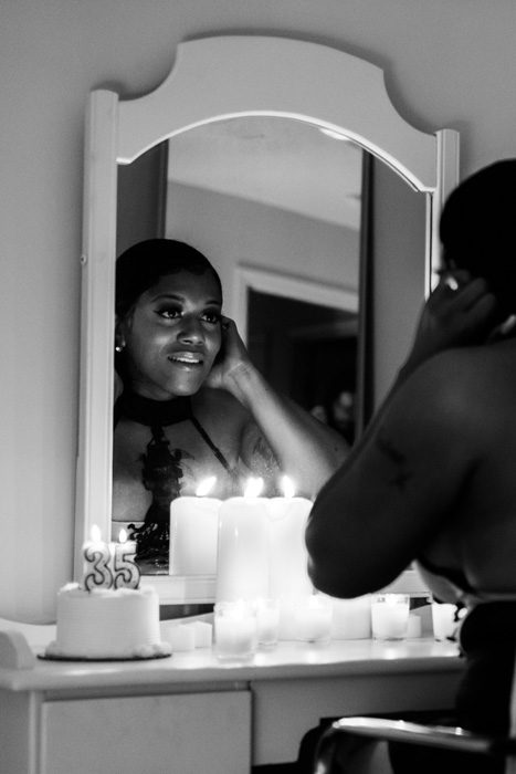 A Black woman sitting in front of a vanity mirror next to a cake that has a burning 35 candle on it.