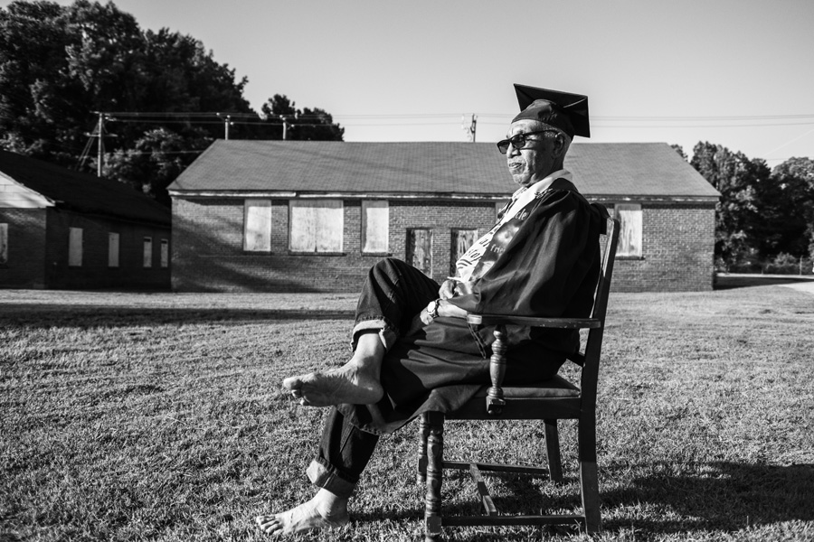 An older Black man sitting in a chair on a lawn in full college graduation regalia.