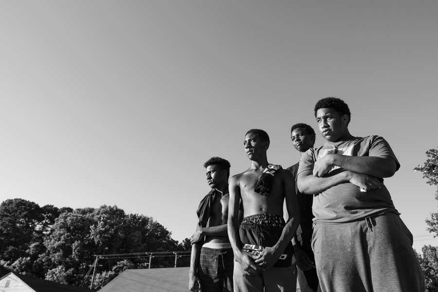 Four Black male teenagers outside in a neighborhood, staring into the middle distance.