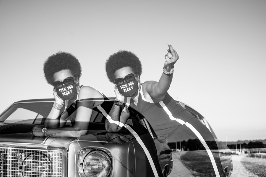 Two Black women superimposed leaning over a car; they wear masks that say "Fuck You, Becky," and one of them is holding up her middle finger. 