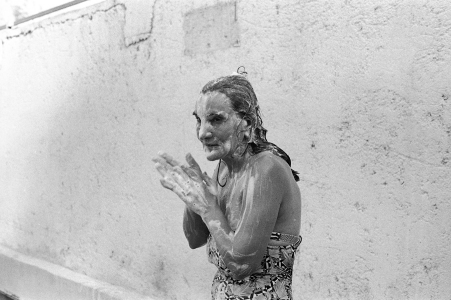 An older woman in a bathing suit, covered in soap suds by the wall of a communal shower.