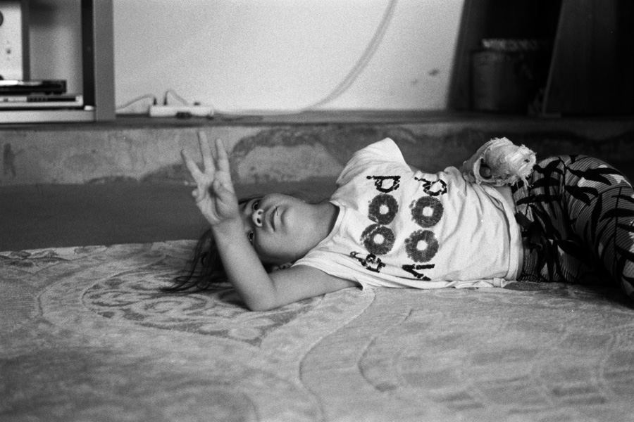 A young girl with her arm in a cast lies on a bed and reaches upward with her good hand.