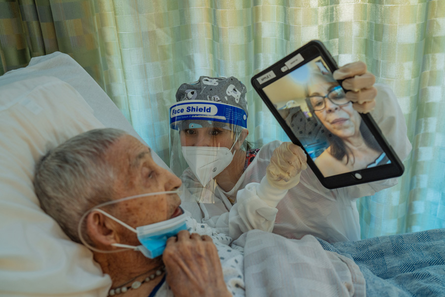 A senior man in a hospital bed talks to a woman in an iPad, held by a nurse in personal protective gear.