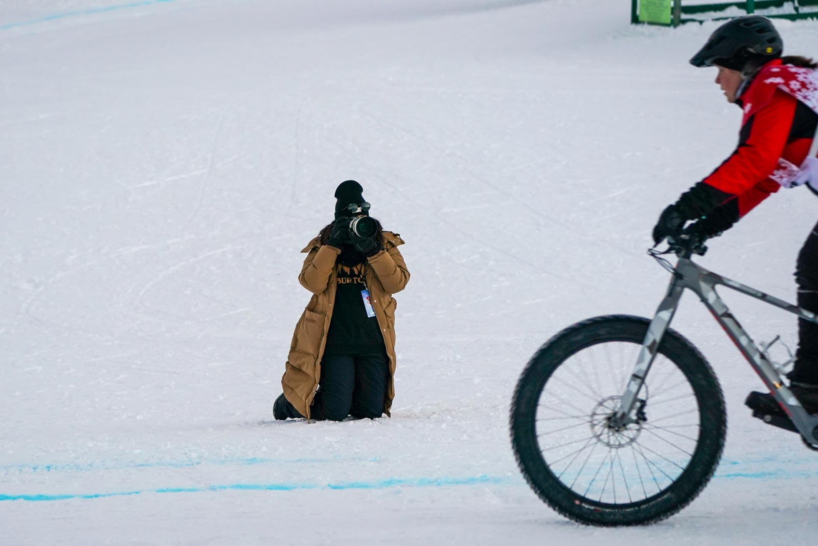 A student kneeling on the snow, taking a picture of a bicyclist racing past.