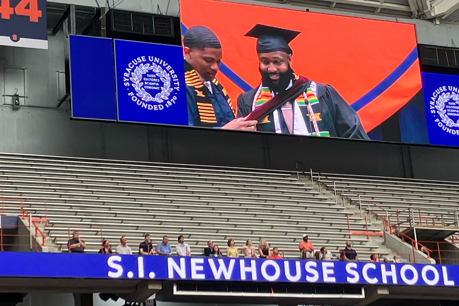 An image on a video board shows a graduate helping to adjust a fellow graduate's academic regalia