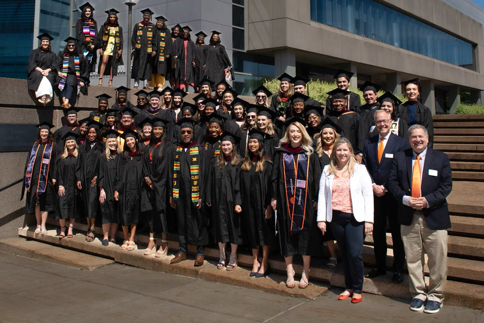 Graduates pose for a photo in academic regalia on the steps of the Newhouse School